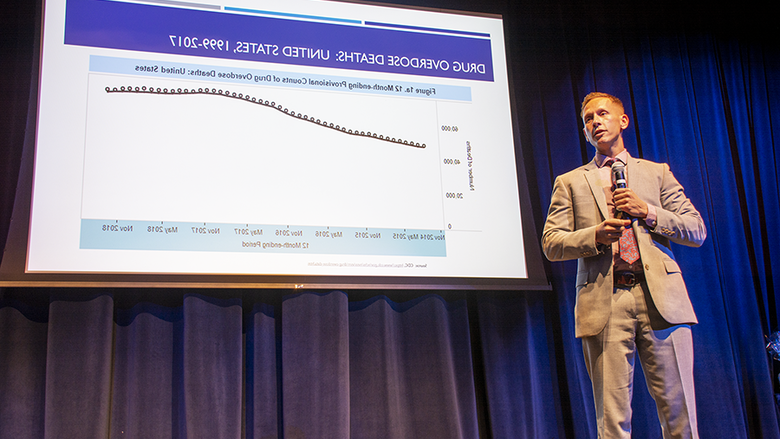 Dr. Glenn Sterner stands on stage as he delivers a speech. There is a slide behind him reviewing drug overdose deaths in the U.S. from 1999-2017 